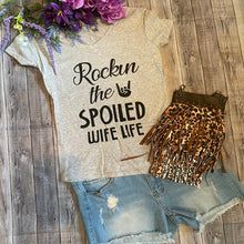 Load image into Gallery viewer, t-shirt with 'Rockin the Spoiled Wife Life' text, leopard print bag, and denim shorts on a wooden surface.